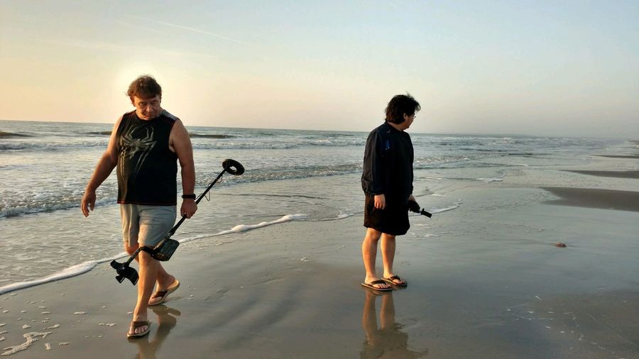 Full length of father and son holding metal detectors at beach against sky