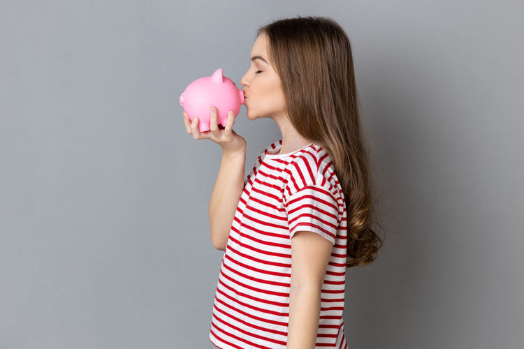 Portrait of smiling young woman holding piggy bank against wall