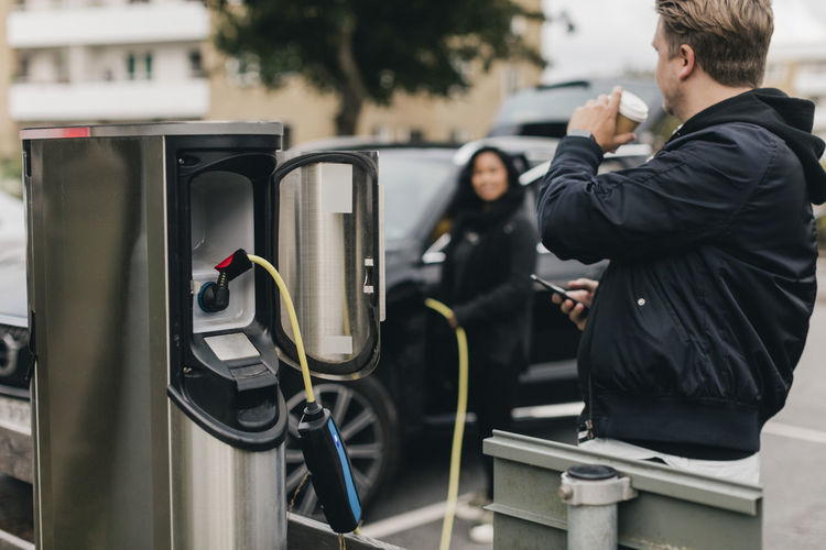 Man drinking coffee while standing at electric car charging station