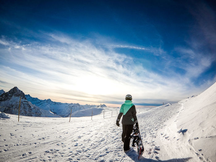 Rear view of man with snowboard walking on snowcapped mountain against sky