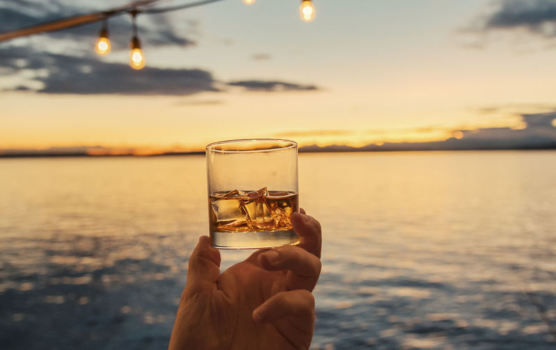 Close-up of hand holding whiskey glass against sea during sunset