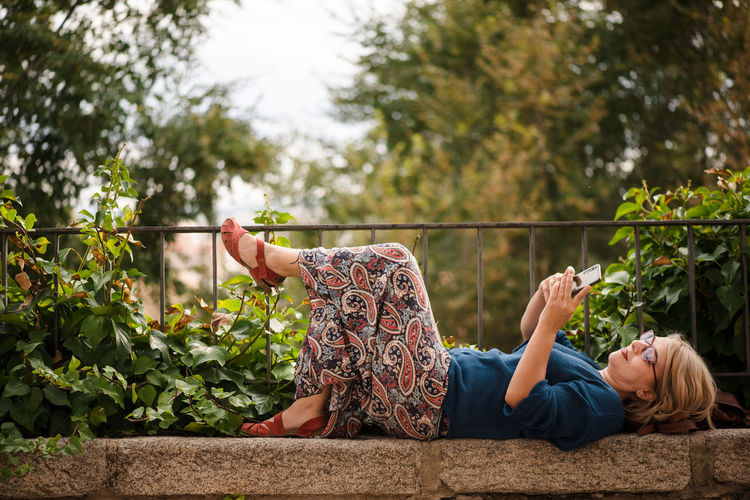 Full body side view of female traveler in protective mask lying on stone border against green trees and browsing mobile phone while relaxing during sightseeing in cuenca town in spain