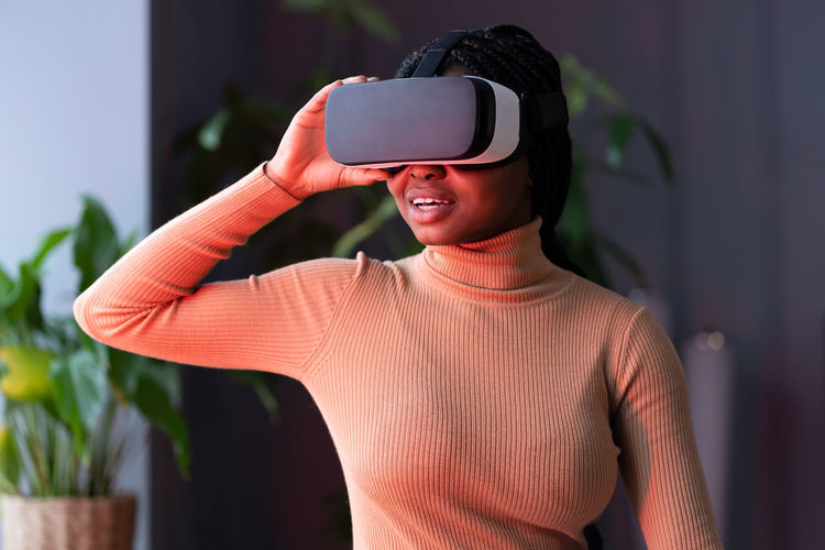 Smiling african woman in vr helmet sitting indoors in red neon light, traveling in 360 degrees