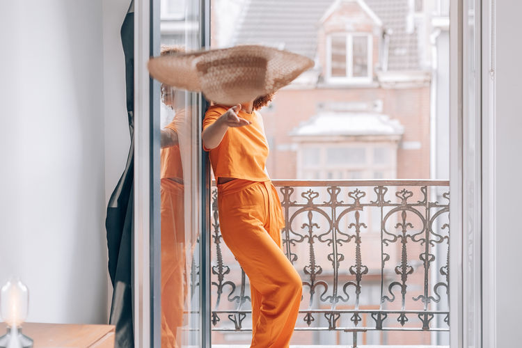 Girl in orange summer suit on the balcony of the room throwing a straw hat on the bed