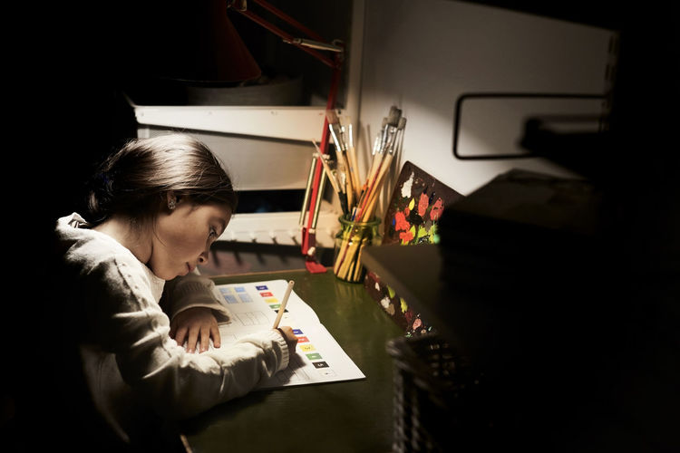 High angle view of girl studying while sitting at illuminated desk in darkroom