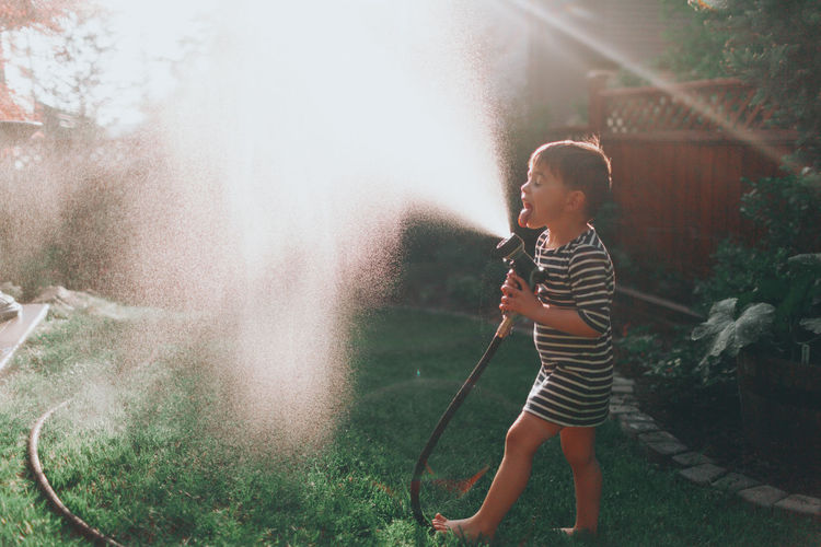 Side view of playful boy sticking out tongue while spraying water through garden hose in lawn