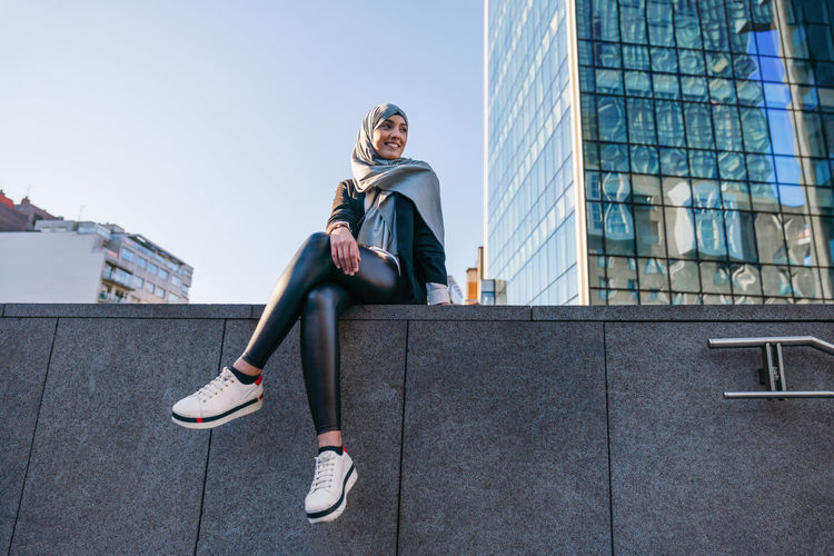 Low angle of smiling muslim female in headscarf and trendy clothes sitting on border in street and looking away