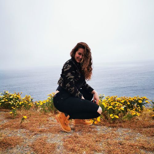 Portrait of smiling woman crouching by plants against sea and sky