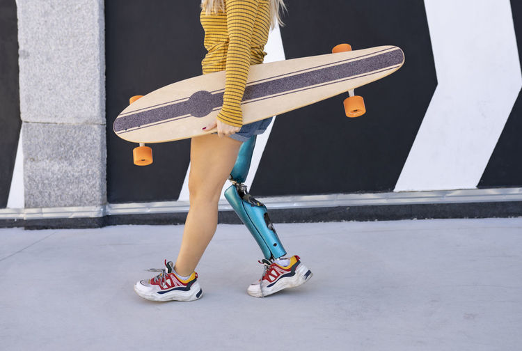 Side view of crop anonymous female with artificial limb of leg walking with skateboard along street in summer