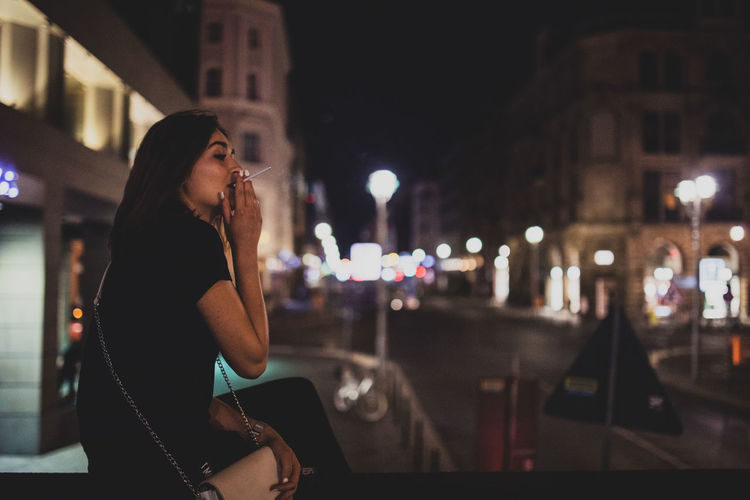 Side view of woman smoking cigarette while sitting in city at night