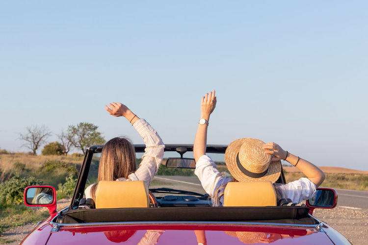 Happy free girlfriends with hat driving in red retro car cheering joyful with arms raised. 