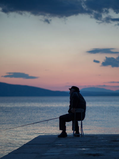 Man sitting on shore against sky during sunset