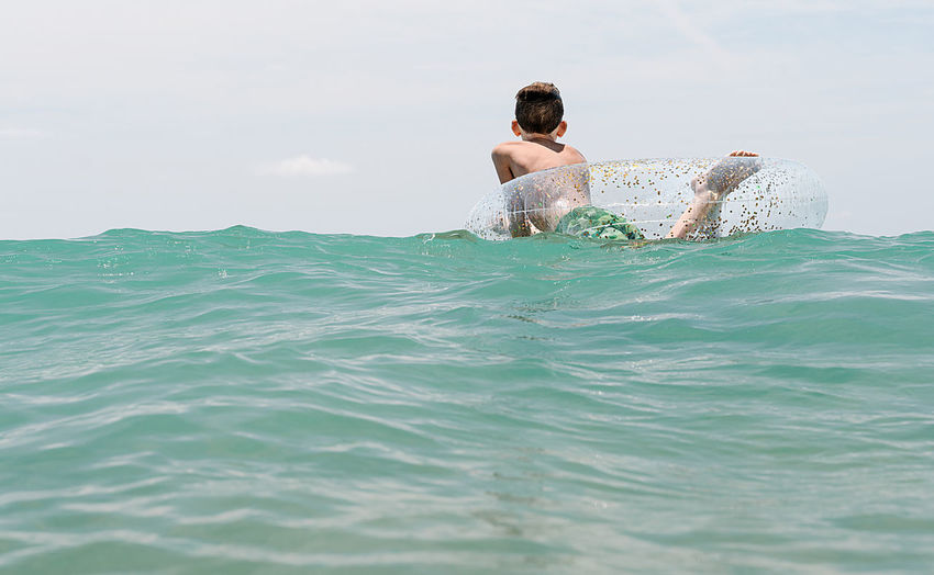 Relaxed white boy swimming in a float in the sea enjoying calm waves in summertime. backwards horizontal photo