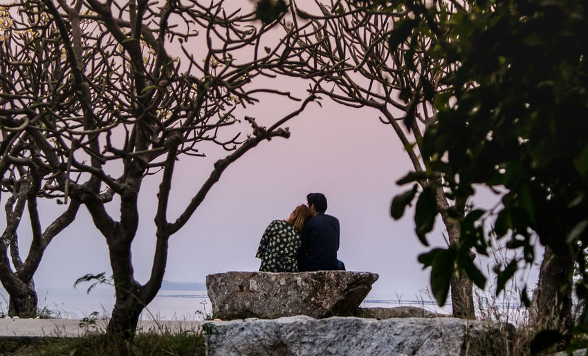 Rear view of couple sitting on rock at beach during sunset