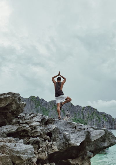 Rear view of man practicing tree pose on rock at beach against cloudy sky