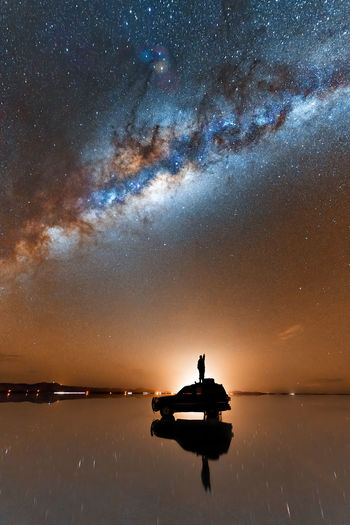 Silhouette of unrecognizable traveler standing on suv car parked on shiny surface of salt flat of salar de uyuni and admiring scenic milky way galaxy shining in evening sky person