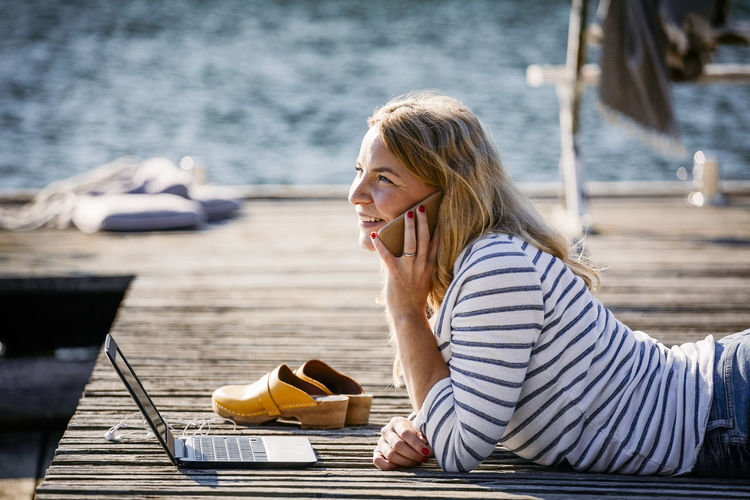 Smiling female with laptop talking on smart phone while lying over wooden pier