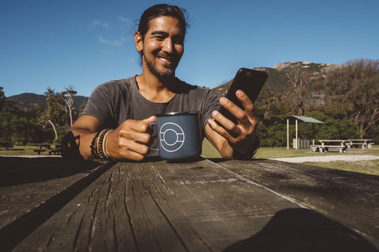 Smiling hiker holding mug while using mobile phone on wooden table at wilsons promontory national park