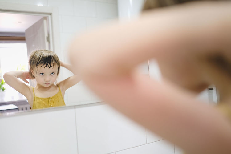 Child in yellow jumpsuit combs his own in front of mirror in bathroom