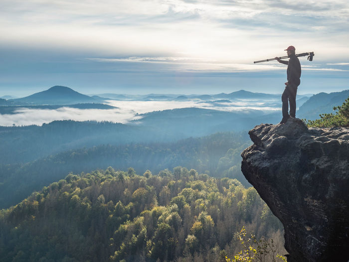 Nature photographer carry camera with tripod on his shoulder and look from mountain cliff to mist