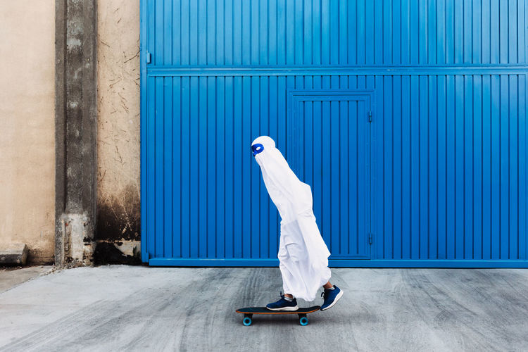 Unrecognizable child wearing mask and white ghost costume standing on skateboard in city during halloween