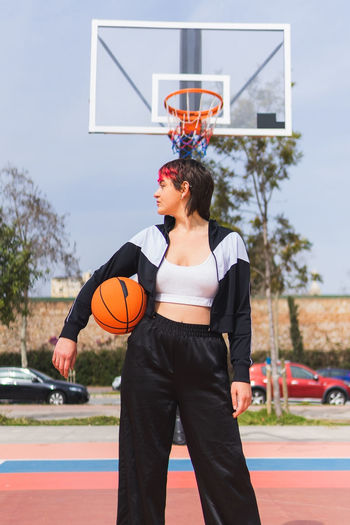 Woman in activewear with ball standing on outdoor sports ground and looking away against backboard with hoop