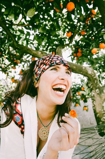 Laughing woman holding flower while standing against tree