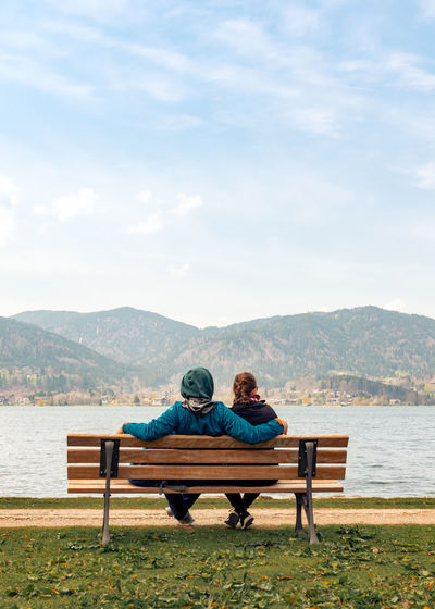 Rear view of man and girl sitting on bench against lake and sky