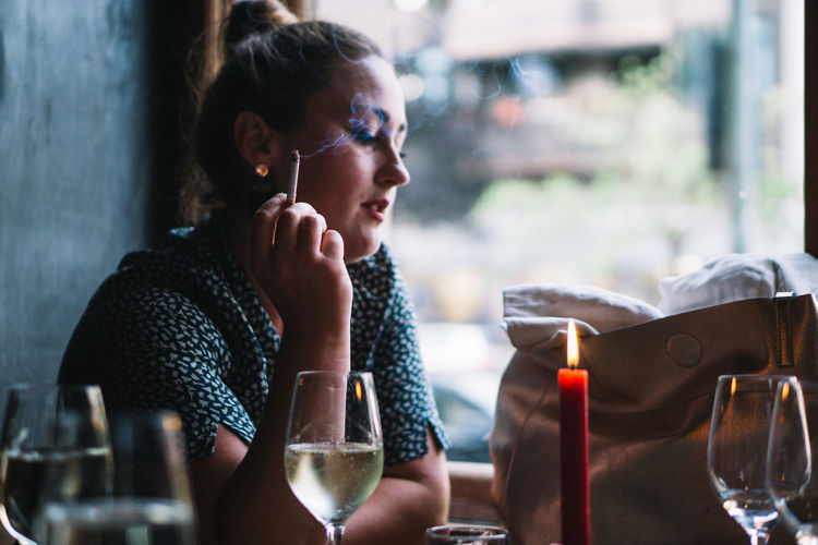 Side view of woman smoking cigarette while sitting with wineglass at restaurant table