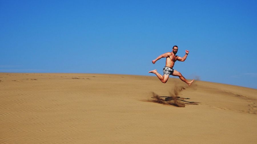 Full length of shirtless man jumping at desert against clear blue sky