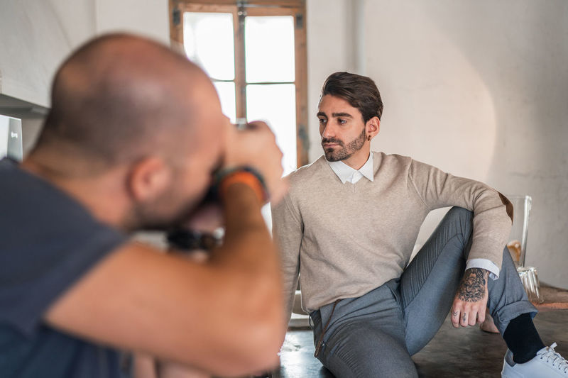 Unrecognizable man focusing and taking shot with camera of stylish adult guy in casual clothes looking away and contemplating while sitting with leg bent on wooden table against small window in light photo studio