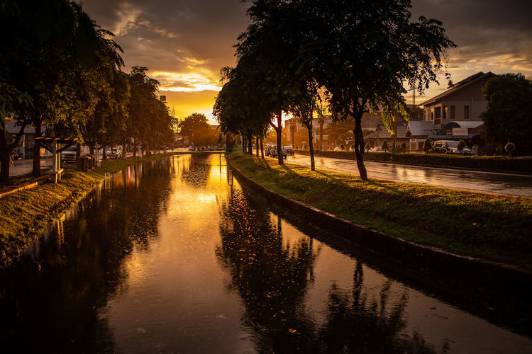 Canal amidst trees and buildings against sky during sunset