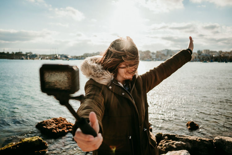 Woman taking selfie with camera while standing against lake