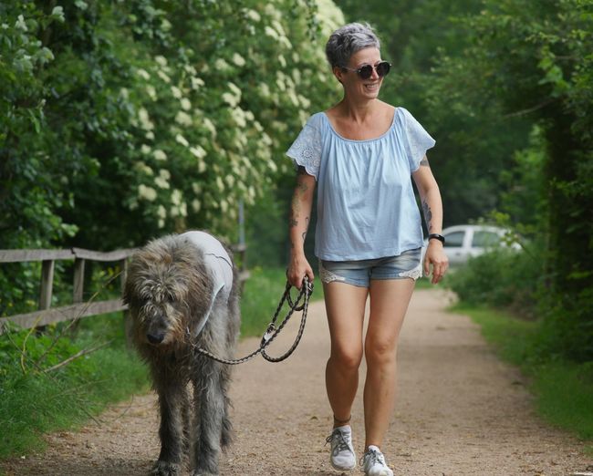 Full length of smiling woman with dog walking on dirt road