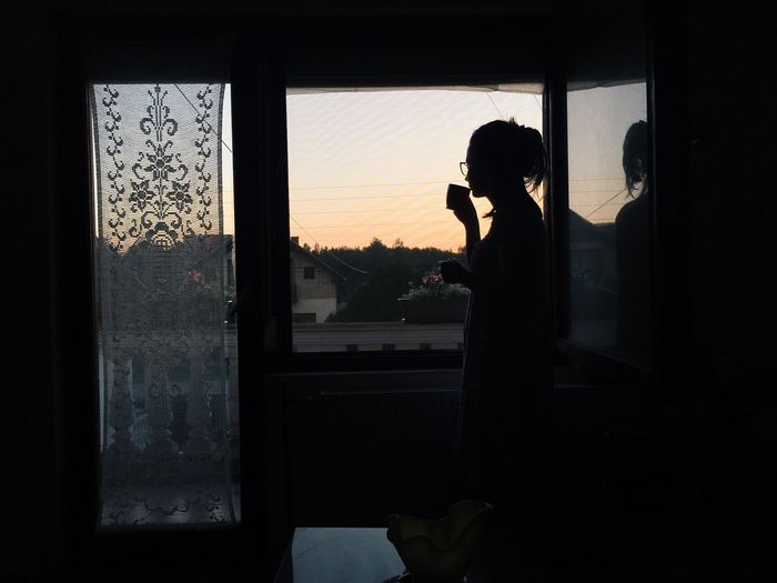 Silhouette woman drinking coffee while standing on window during sunset