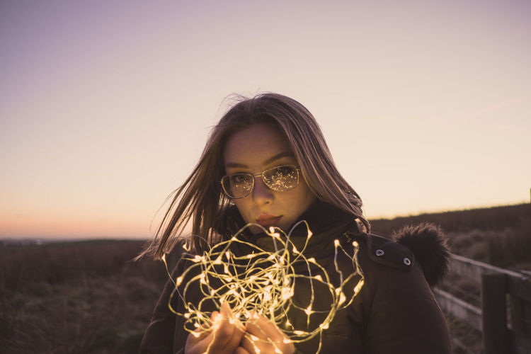 Portrait of woman holding illuminated string lights on field against sky during sunset