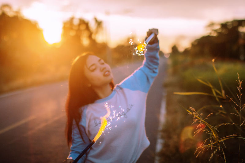 Digital composite image of woman holding firework on road during sunset