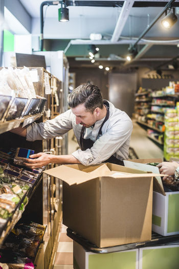 Salesman unloading cardboard box while arranging packets on shelf in store