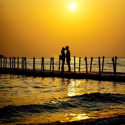 Silhouette couple kissing while standing on pier over sea against orange sky during sunset