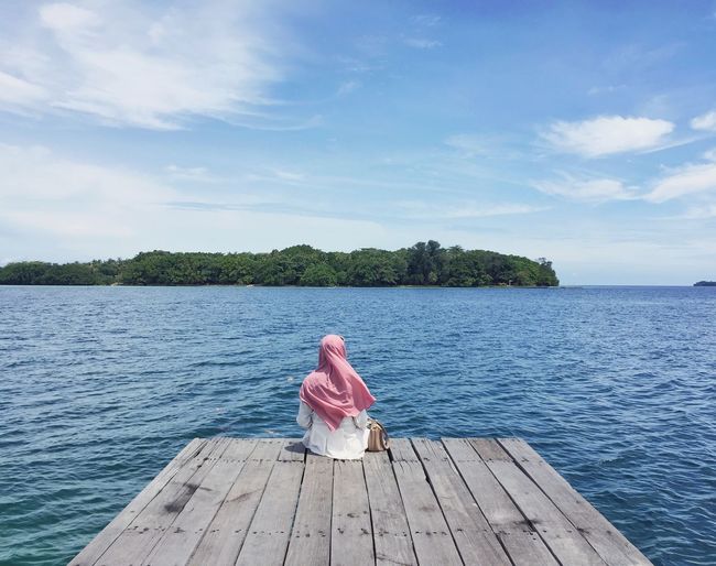 Rear view of young woman sitting on pier over lake against sky