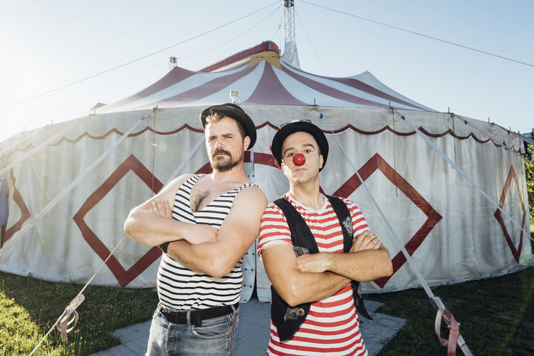 Muscular build artist standing with arms crossed by clown in front of circus tent