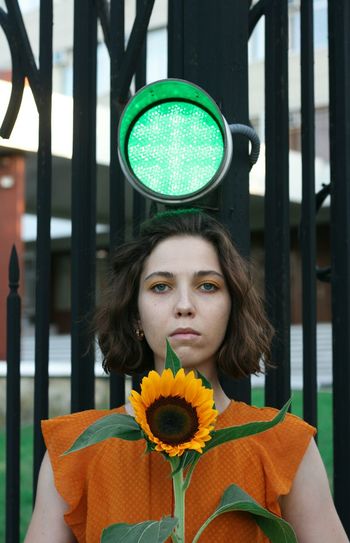 Portrait of serious woman holding sunflower standing against gate