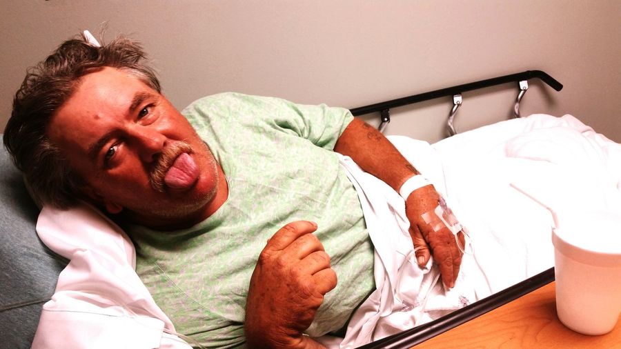 High angle portrait of man sticking out tongue while sitting on bed in hospital