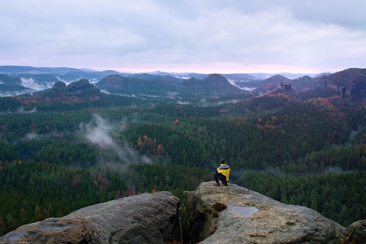 Rear view of male hiker in yellow black jacket on rocky peak while enjoying a daybreak above valley