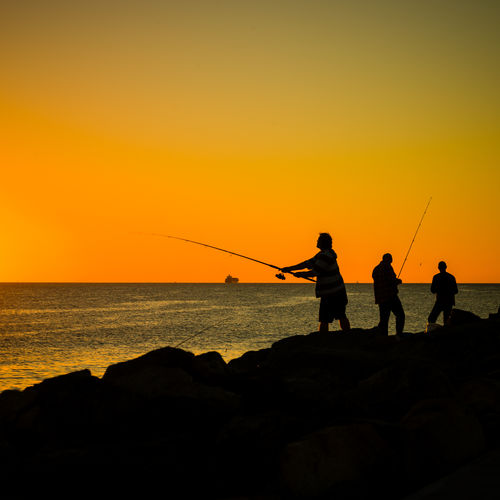 Silhouette people fishing in sea against clear orange sky