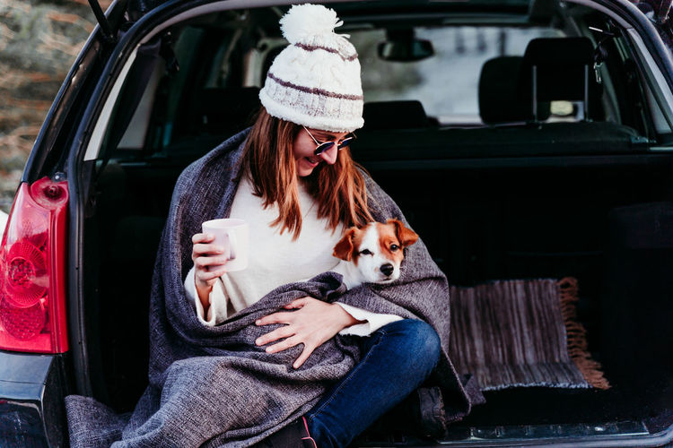 Woman holding mug sitting with dog in car trunk