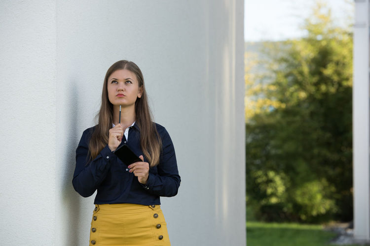 Thoughtful woman with mobile phone standing by concrete wall