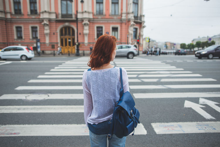 Back view of anonymous female with red hair standing near crosswalk in saint petersburg and looking away