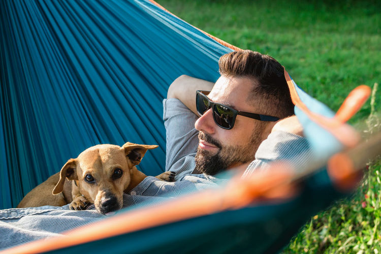 Smiling attractive european man in sunglasses is resting in hammock with his cute little dog.