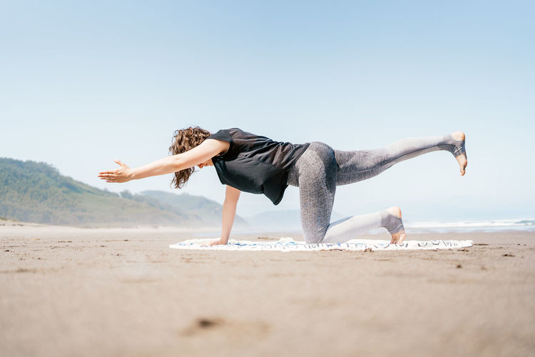 Side full body young female in sportswear practicing yoga on sandy seashore with eyes closed
